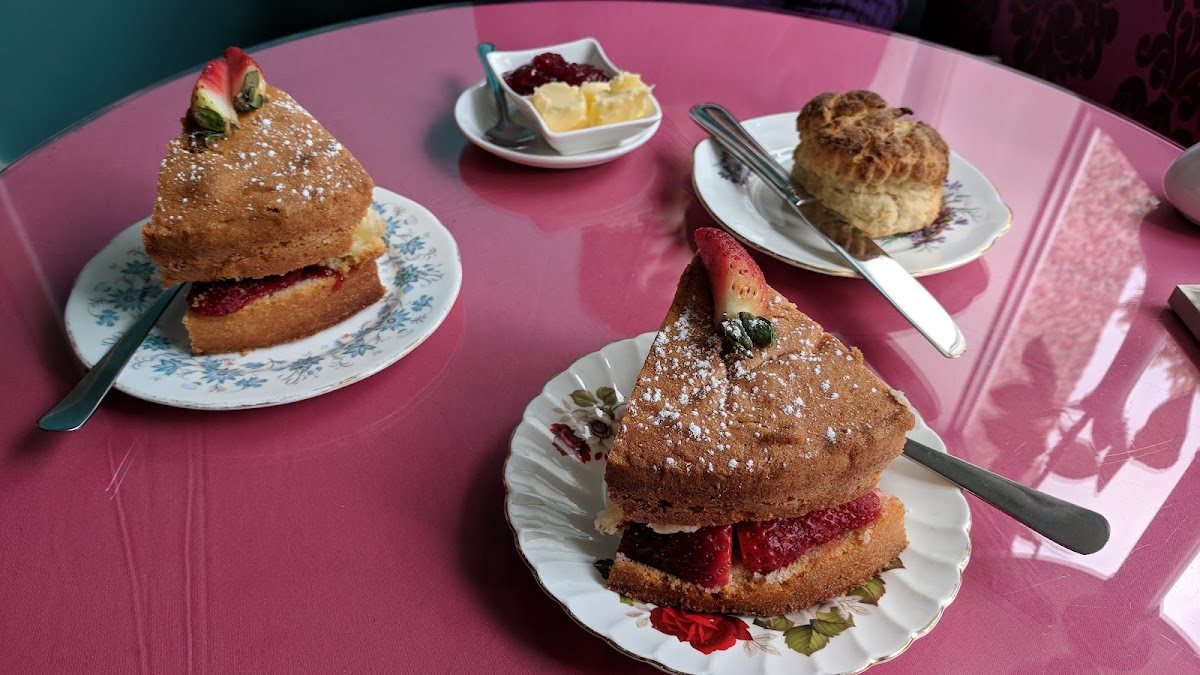 Two tall wedges of Victoria sponge filled with jam and strawberries, dusted with sugar and topped with a strawberry, and a scone with jam and butter, on vintage floral plates on a rose-pink table.