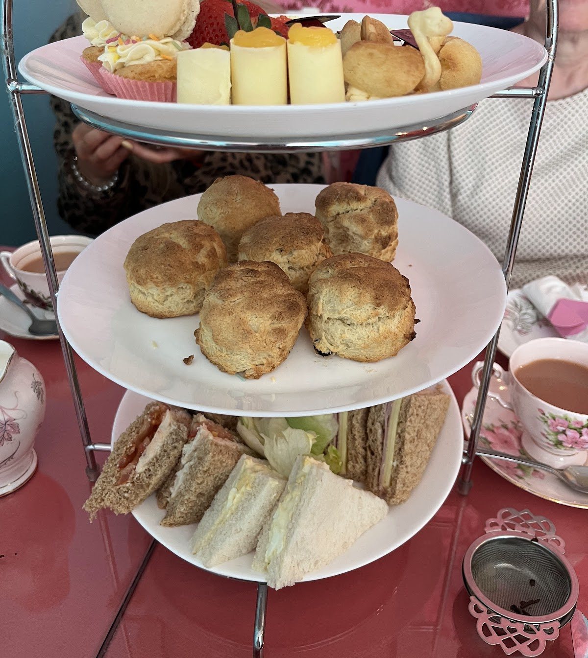 A three-tier afternoon tea stand: macarons, strawberries, lemon curd rolls and choux on top; golden scones in the middle; finger sandwiches on the bottom; with a floral teacup and a rose-pink table.