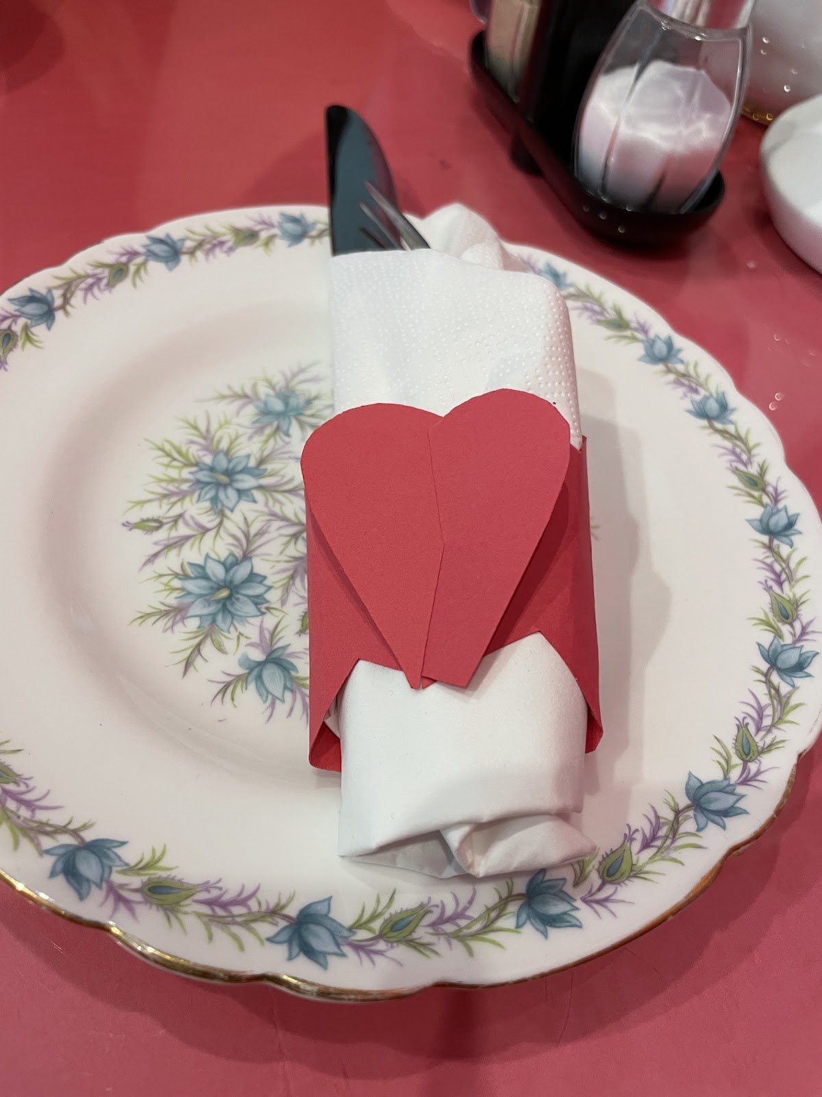 A white napkin wrapped in a red paper heart on a vintage plate with a blue floral border.