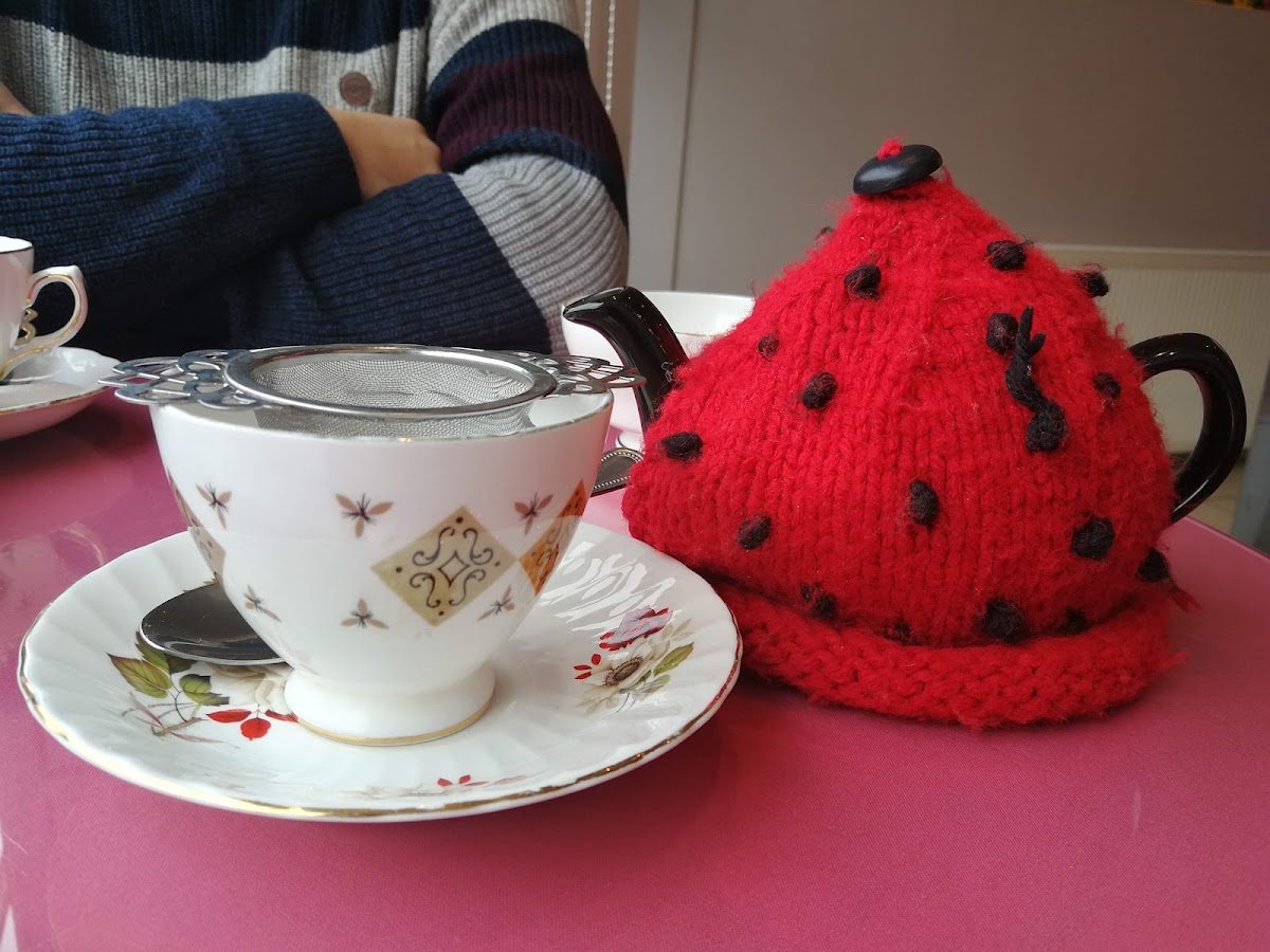 A red knitted tea cosy with black spots like a ladybird over a teapot, next to a bone-china cup and saucer with a silver tea strainer, on a rose-pink table.